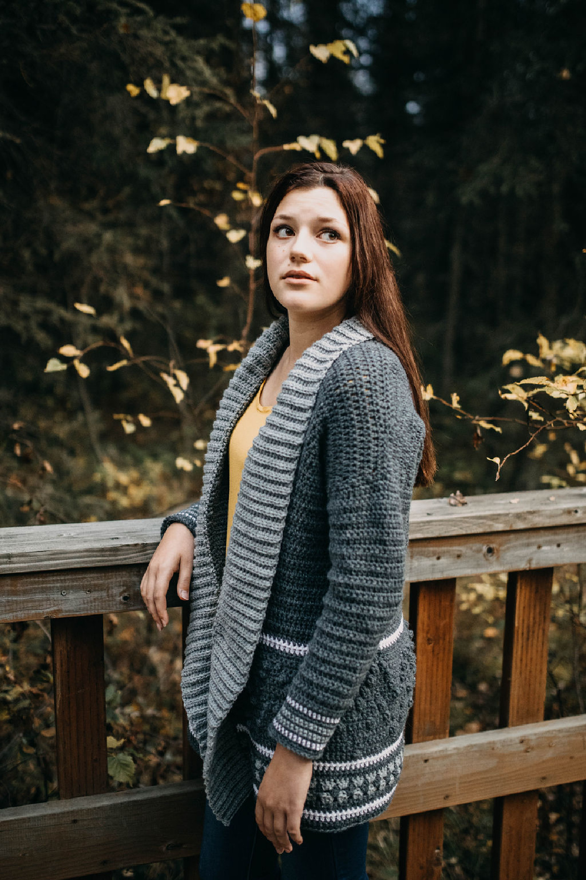 Woman wearing a grey crochet cardigan and a yellow top with her arm on a wooden fence.