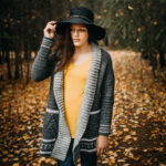 Woman wearing a grey crochet cardigan with a yellow top with her hand on the brim of a black hat.