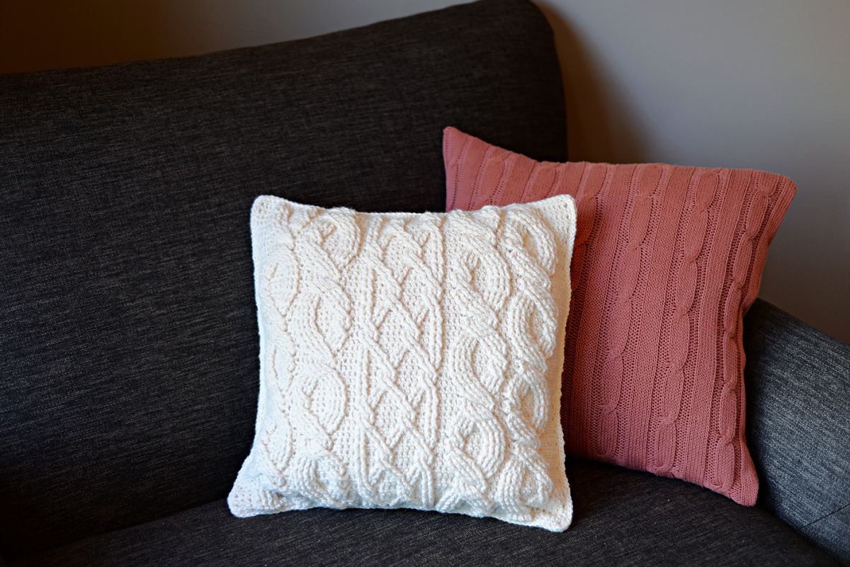 White cabled crochet pillow and pink cushion on a dark sofa.