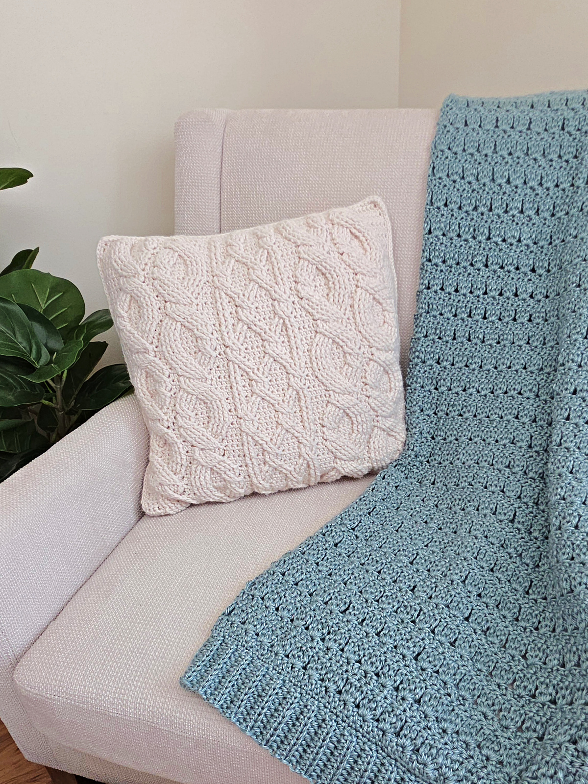 White cabled crochet pillow on a sofa with a light blue crochet blanket and plant next to it.