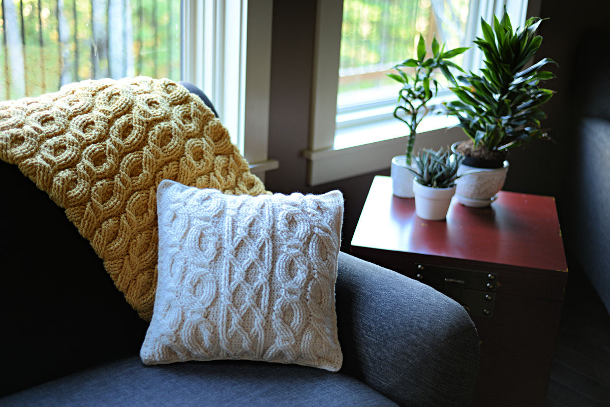 White cabled crochet pillow on a sofa with a yellow crochet blanket behind it and a plant next to it.