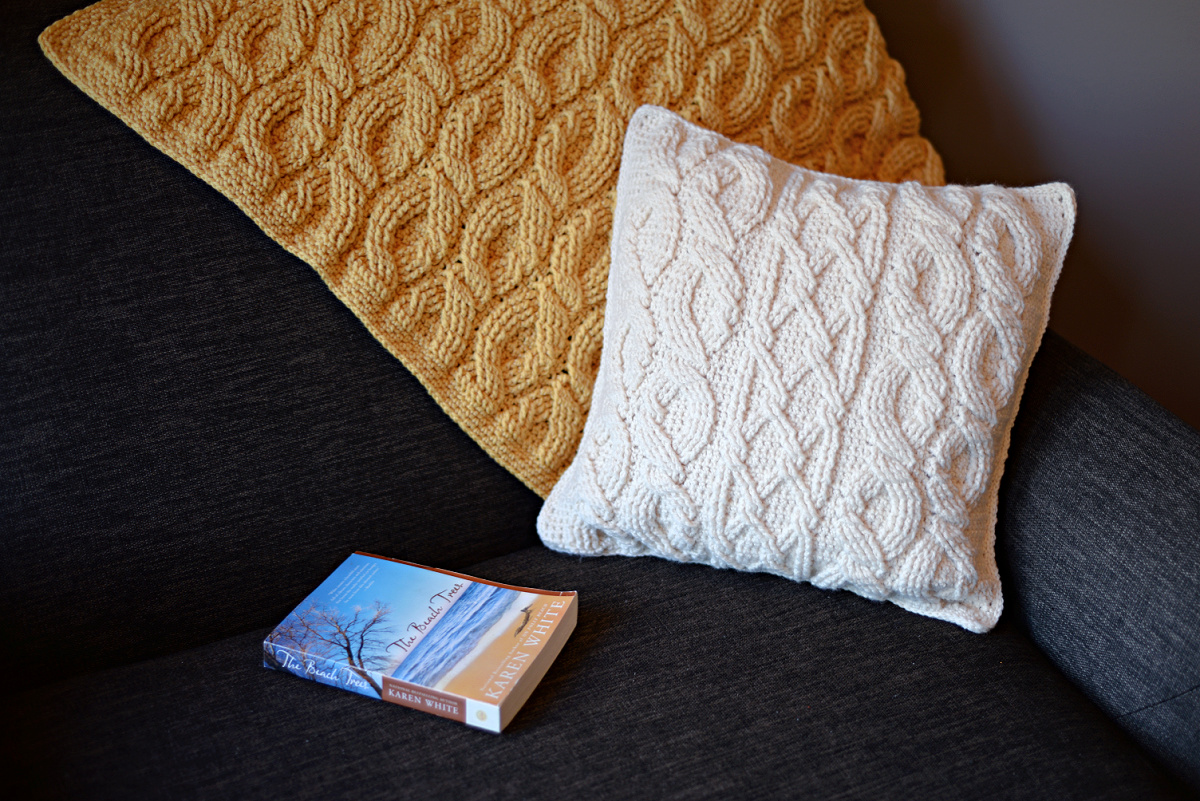 White cabled crochet pillow on a sofa with a yellow crochet blanket behind it and a book next to it.