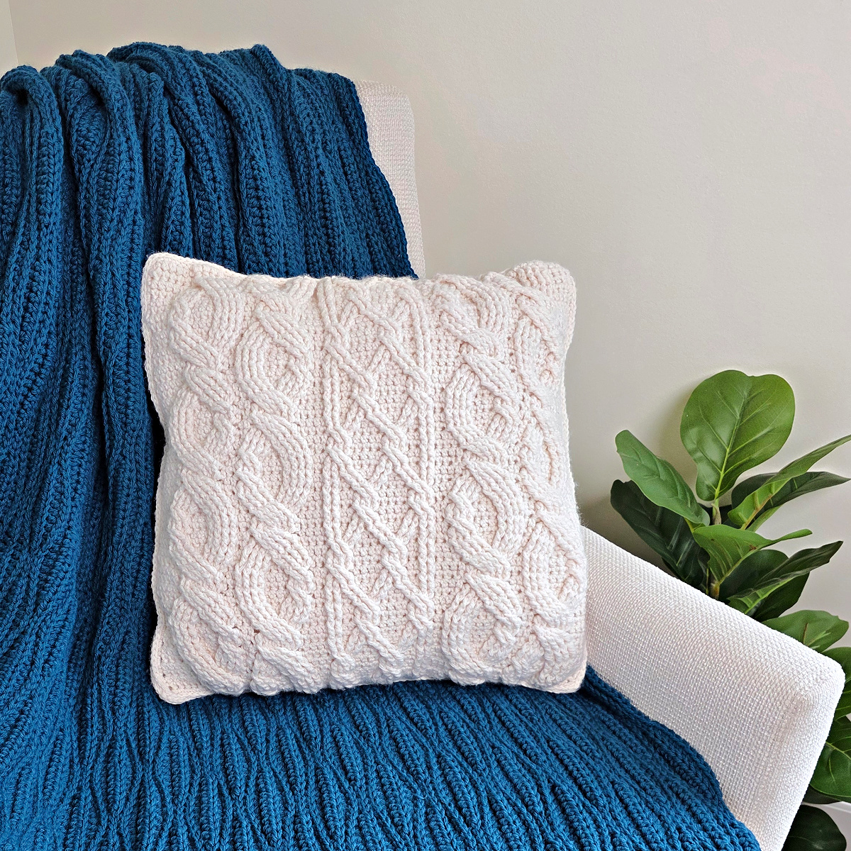 White cabled crochet pillow on a sofa with a blue crochet blanket behind it and a plant next to it.