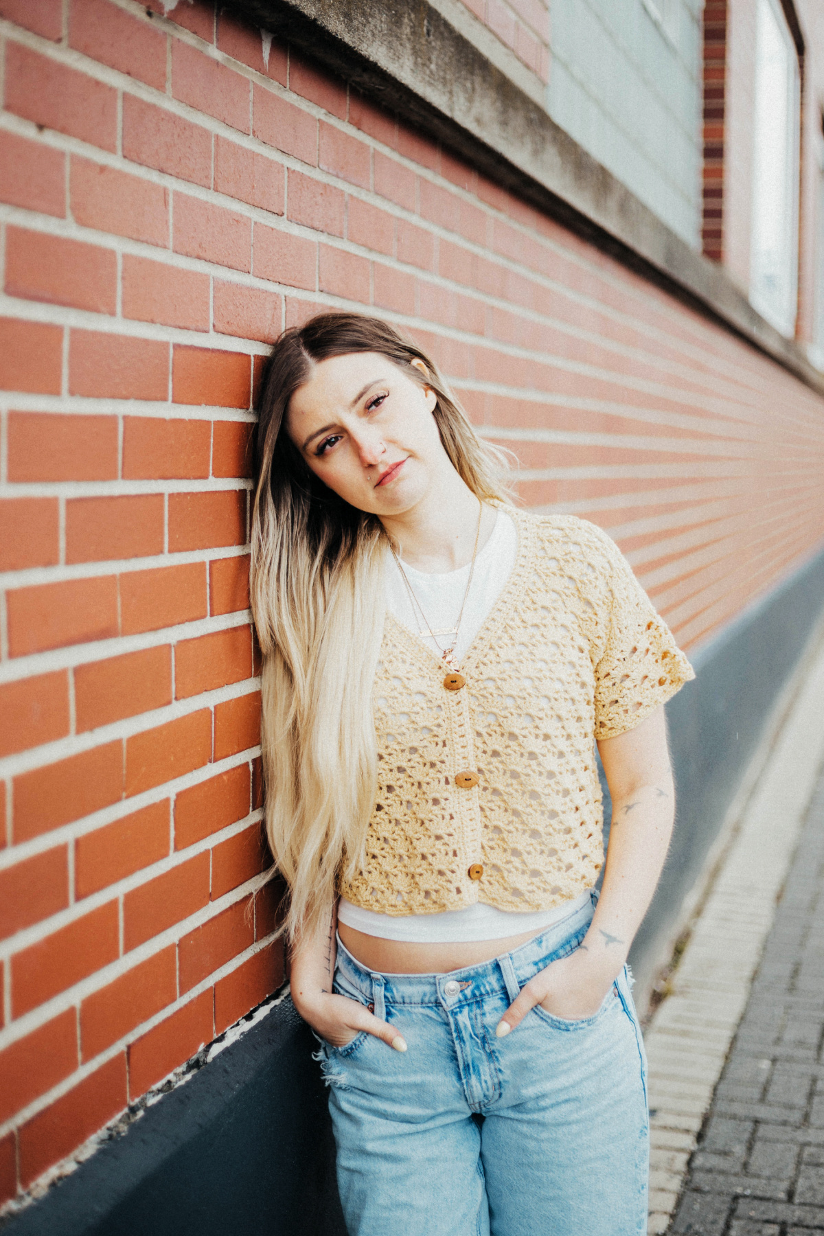 Girl wearing a beige colored cropped crochet cardigan and blue jeans leaning against a brick wall.