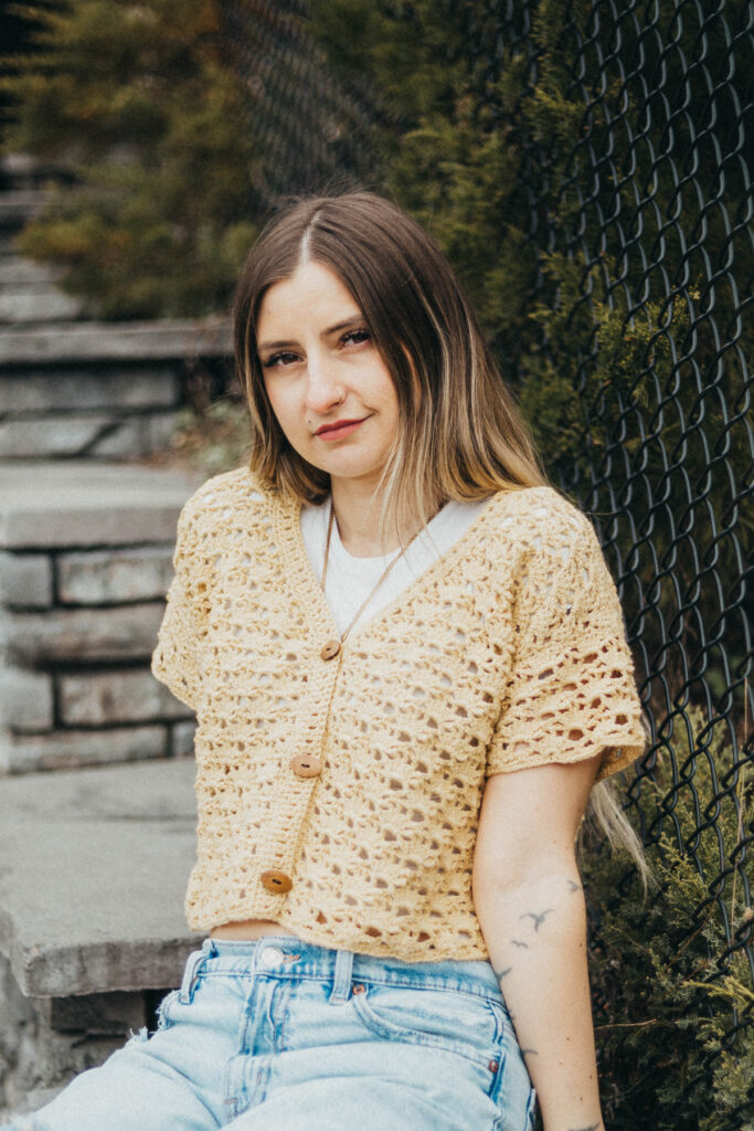 Girl wearing a beige colored cropped crochet cardigan and blue jeans sitting against a stone wall and fence.
