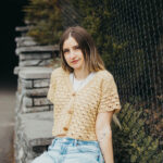 Girl wearing a beige colored cropped crochet cardigan and blue jeans sitting against a stone wall and fence.