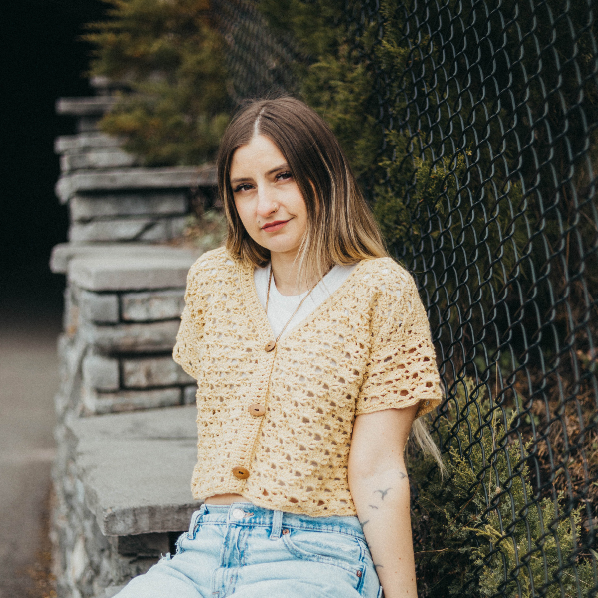 Girl wearing a beige colored cropped crochet cardigan and blue jeans sitting against a stone wall and fence.