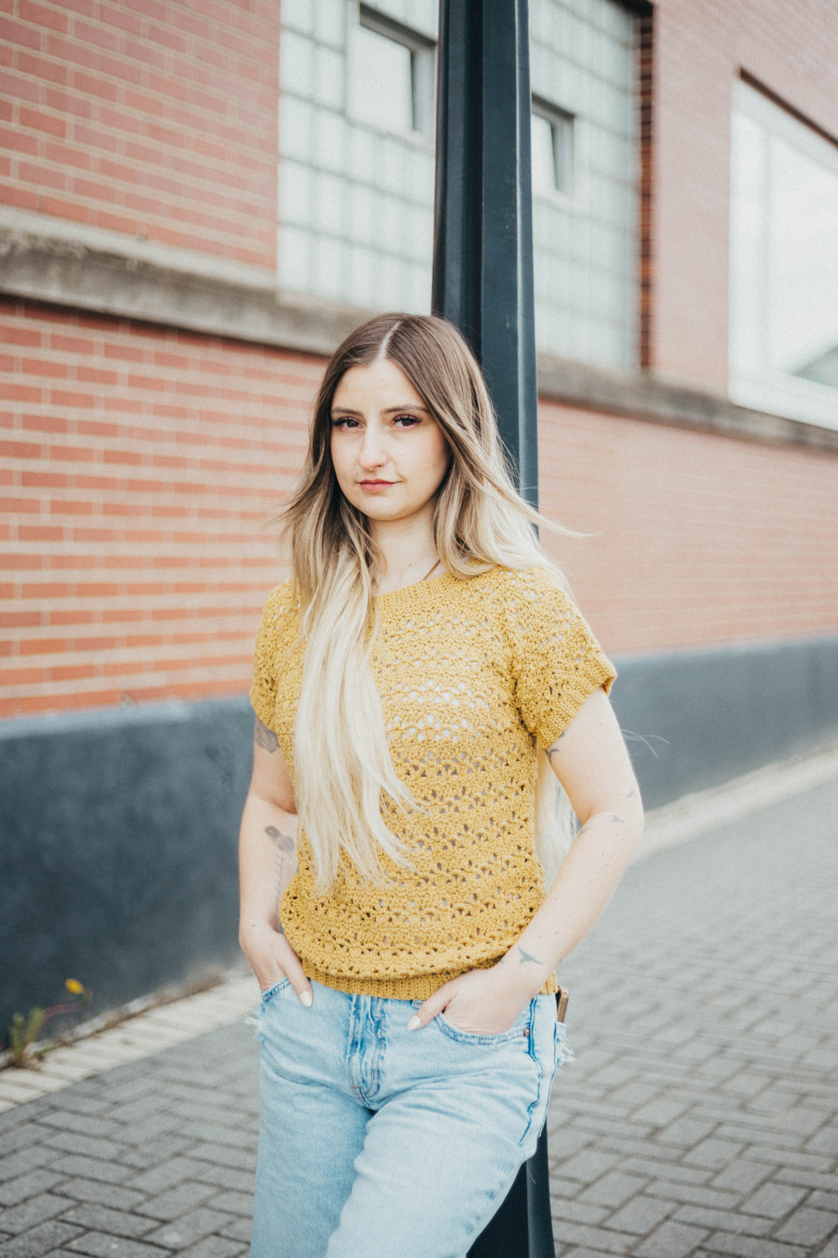 Girl leaning against a pole wearing a summer crochet tee with blue jeans and her hands tucked in her pockets.