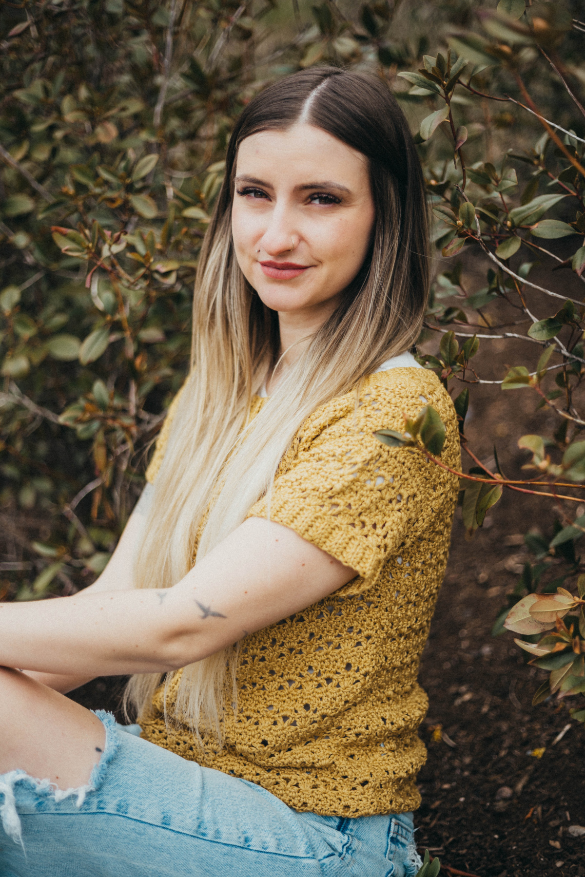 Girl wearing a summer crochet tee with blue jeans sitting against a wall of leaves.