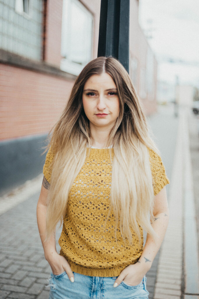 Girl leaning against a pole wearing a summer crochet tee with blue jeans and her hands tucked in her pockets.