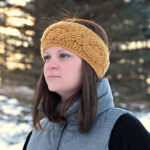 A young girl with red hair wearing a crochet ear warmer outside in the snowy weather.