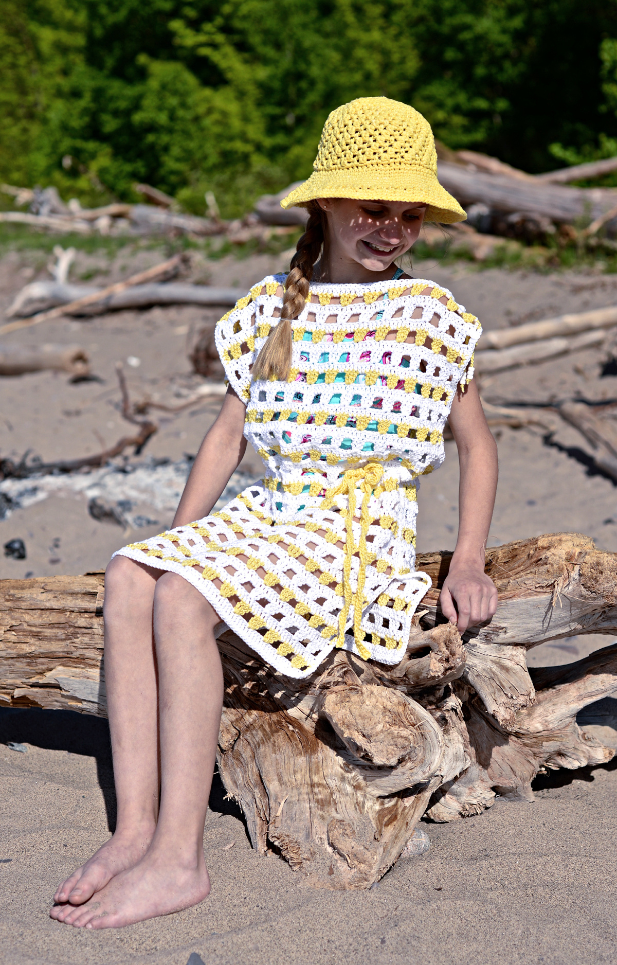 Girl modeling a white and yellow crochet beach cover up and yellow sunhat while sitting on a log at the beach.