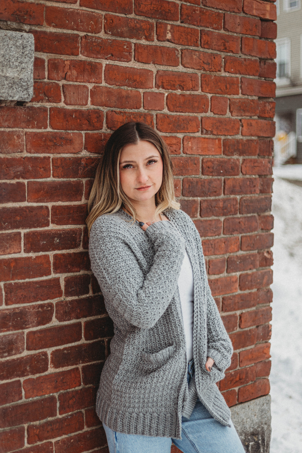 Blonde haired girl wearing a grey crochet cardigan with a white top and jeans leaning against a brick wall.