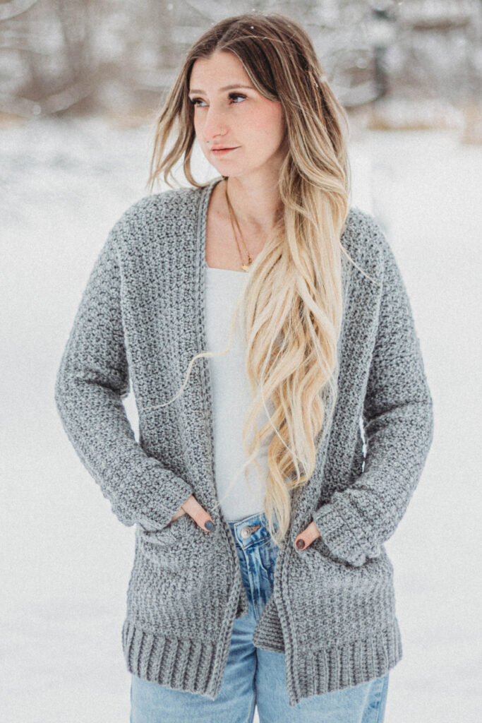 Blonde haired girl wearing a grey crochet cardigan with a white top and jeans.