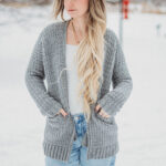 Blonde haired girl wearing a grey crochet cardigan with a white top and jeans.