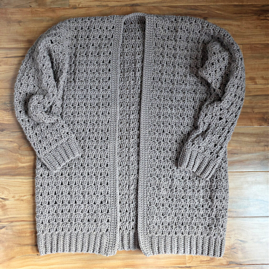 A taupe colored crochet cardigan sweater pattern completed and laying flat on a wood floor.