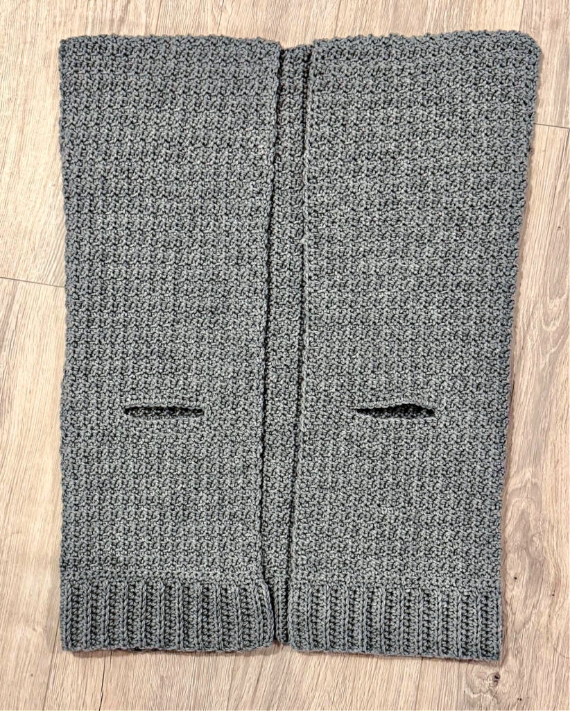A grey crochet cardigan in progress against a wooden background.