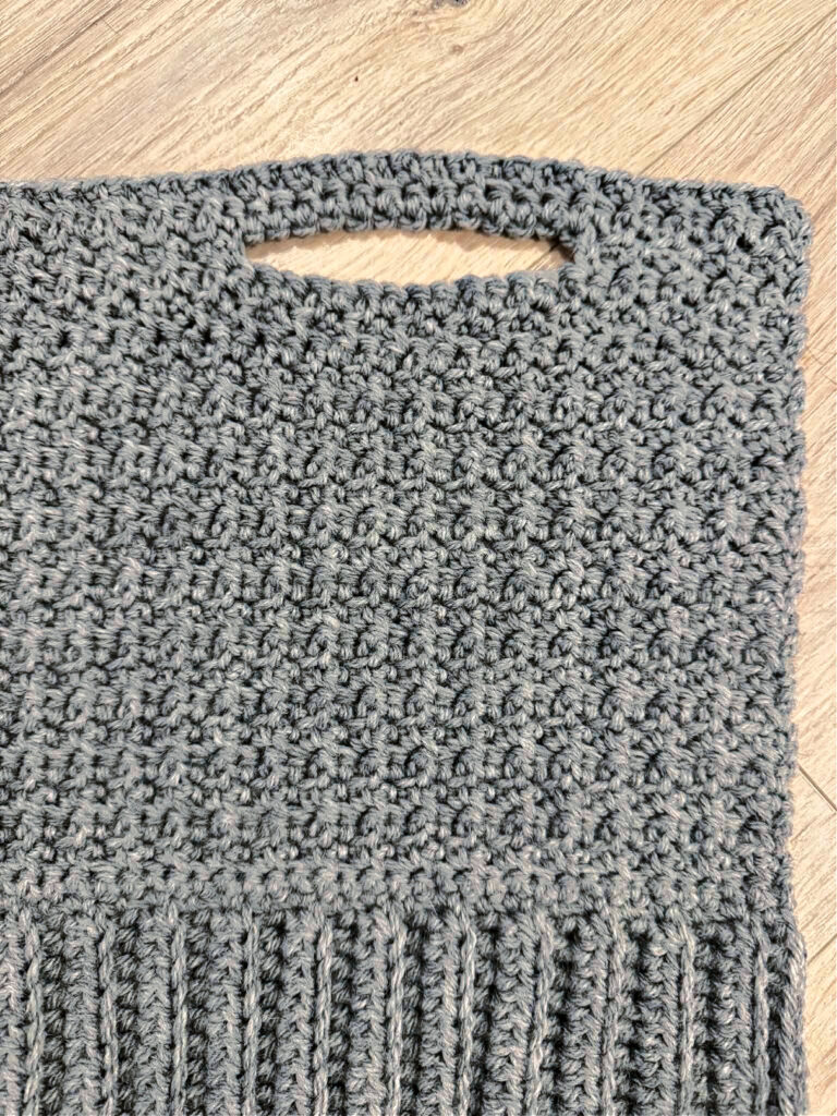 A grey crochet cardigan in progress against a wooden background.