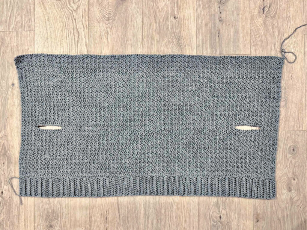 A grey crochet cardigan in progress against a wooden background.