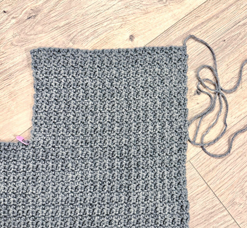 A grey crochet cardigan in progress against a wooden background.
