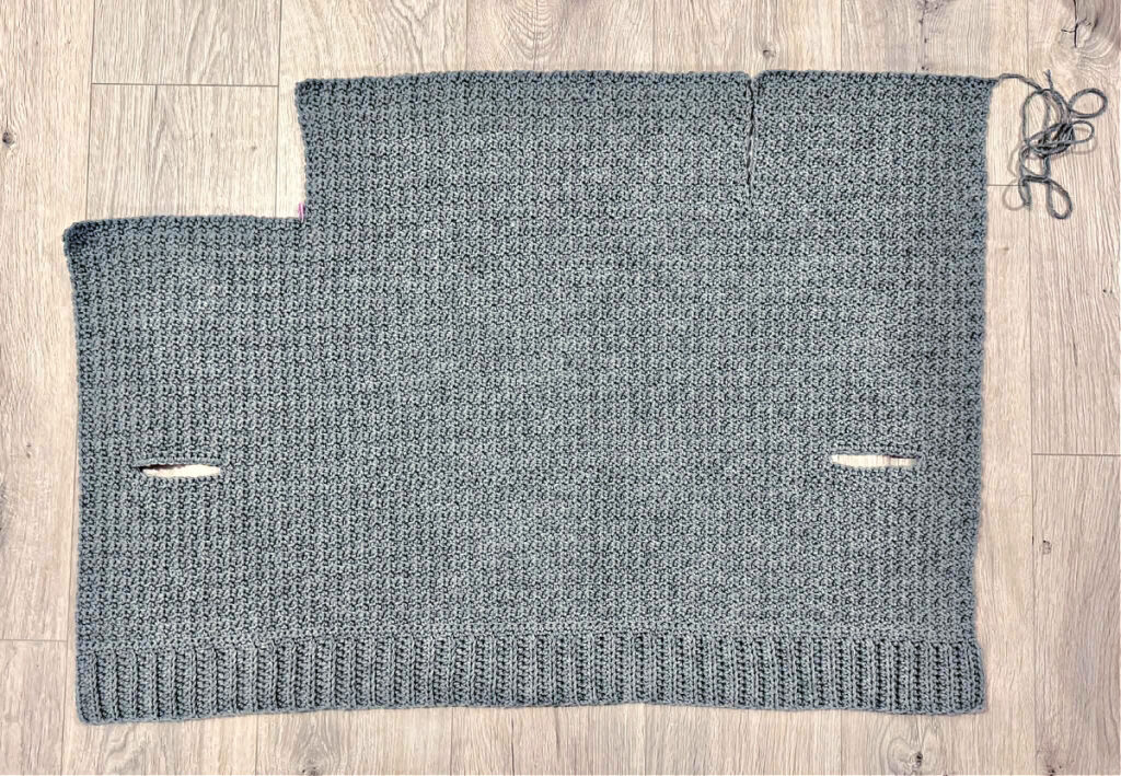A grey crochet cardigan in progress against a wooden background.