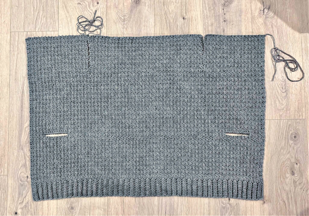 A grey crochet cardigan in progress against a wooden background.