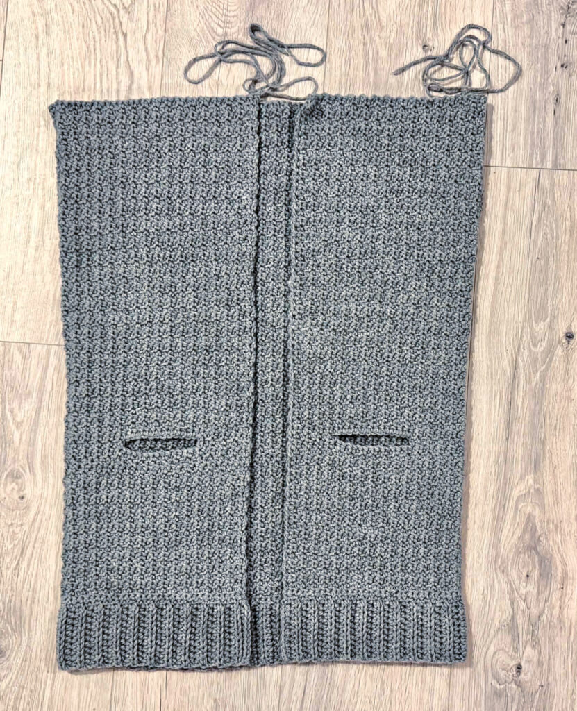 A grey crochet cardigan in progress against a wooden background.