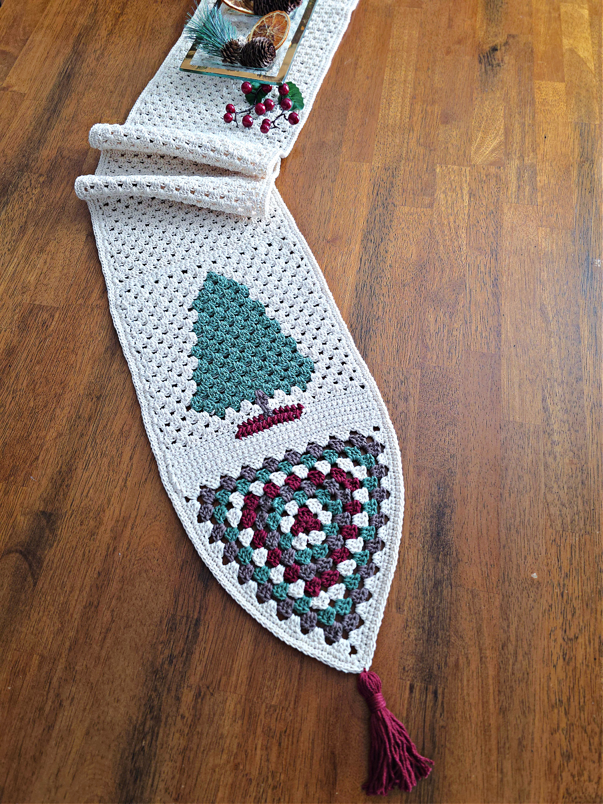 Crochet table runner on a wooden table with a fir tree and granny triangle at one end.