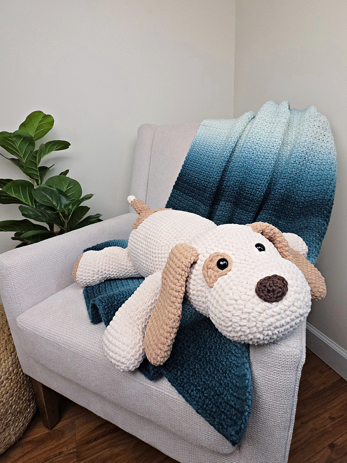 A large plush white and brown crochet dog on a sofa with a blue crocheted blanket thrown over it.