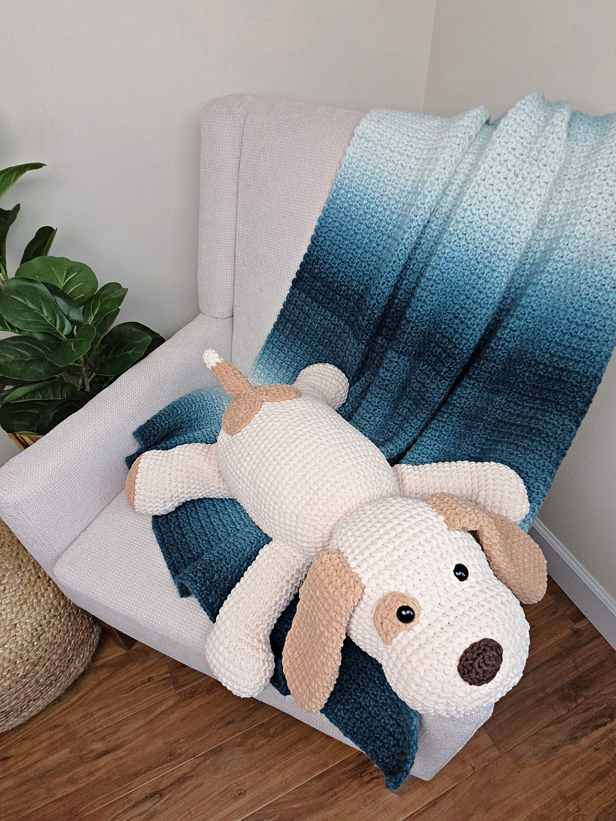 A large plush white and brown crochet dog on a sofa with a blue crocheted blanket thrown over it.