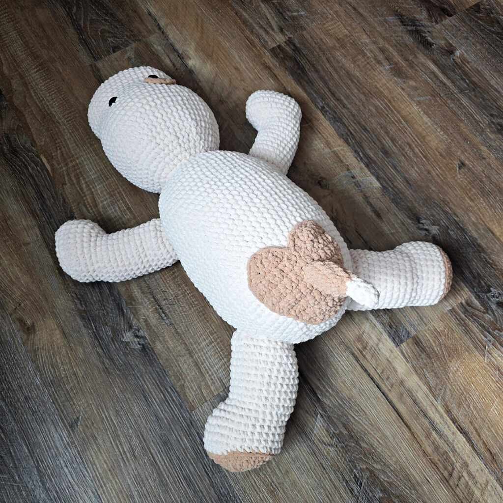 A large plush white and brown crochet dog with floppy ears and a short tail on a wooden floor.