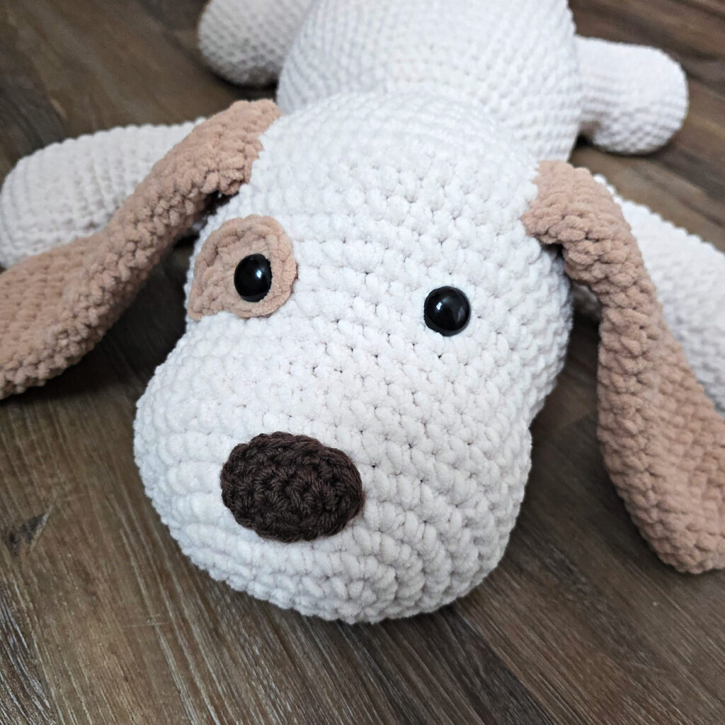 A large plush white and brown crochet dog with floppy ears on a wooden floor.