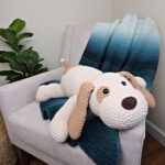 A large plush white and brown crochet dog on a sofa with a blue crocheted blanket thrown over it.