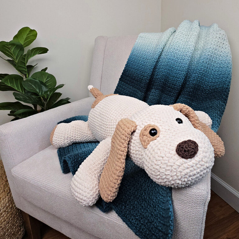 A large plush white and brown crochet dog on a sofa with a blue crocheted blanket thrown over it.