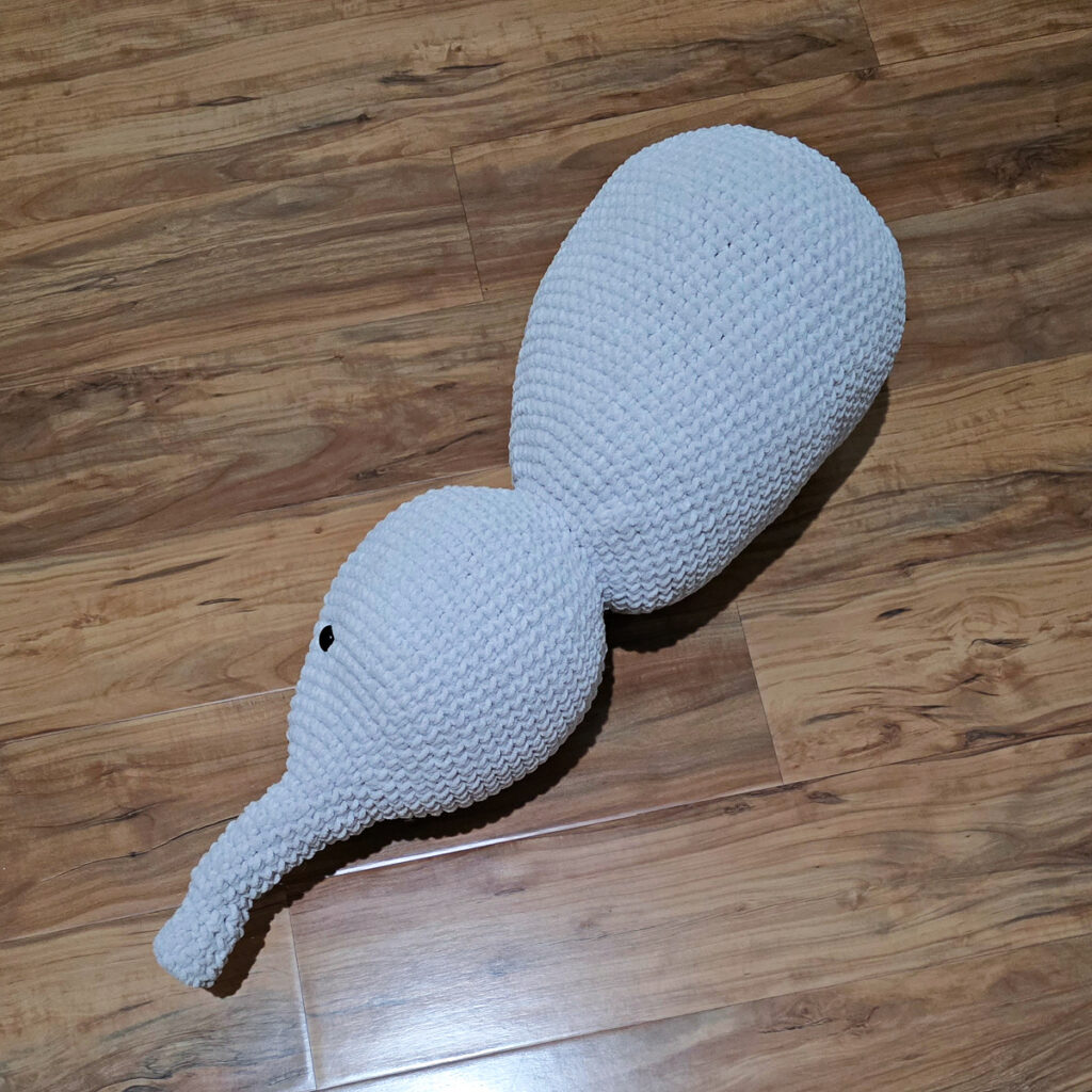 A gray crochet elephant head and body in progress on a wooden floor.