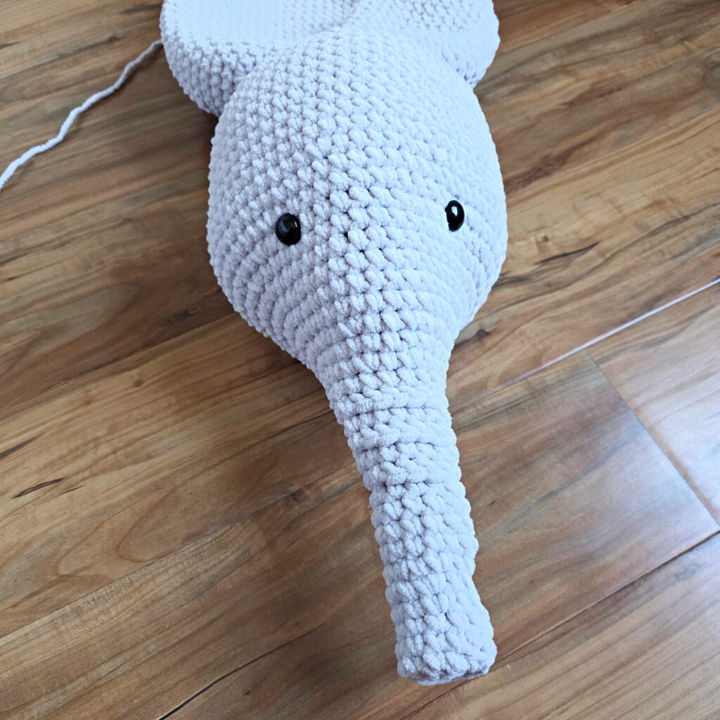 A gray crochet elephant head in progress on a wooden floor.