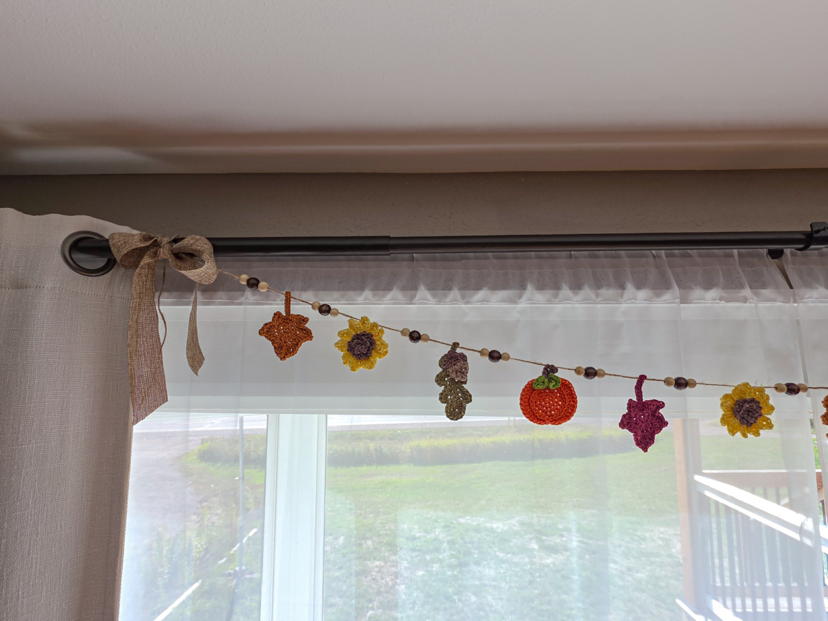A colorful autumn harvest crochet garland with sunflowers, pumpkins, and leaves hung up on the window.