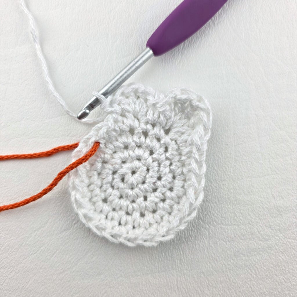 A white crochet ghost with a purple hook and green yarn to mark the stitch.