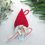 A crochet crochet gnome candy cane holder with a red santa hat next to green tinsel.