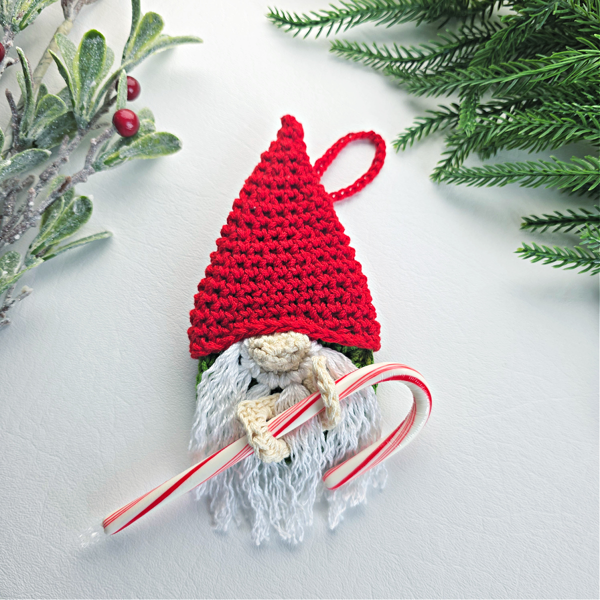 A crochet crochet gnome candy cane holder with a red santa hat next to green tinsel.