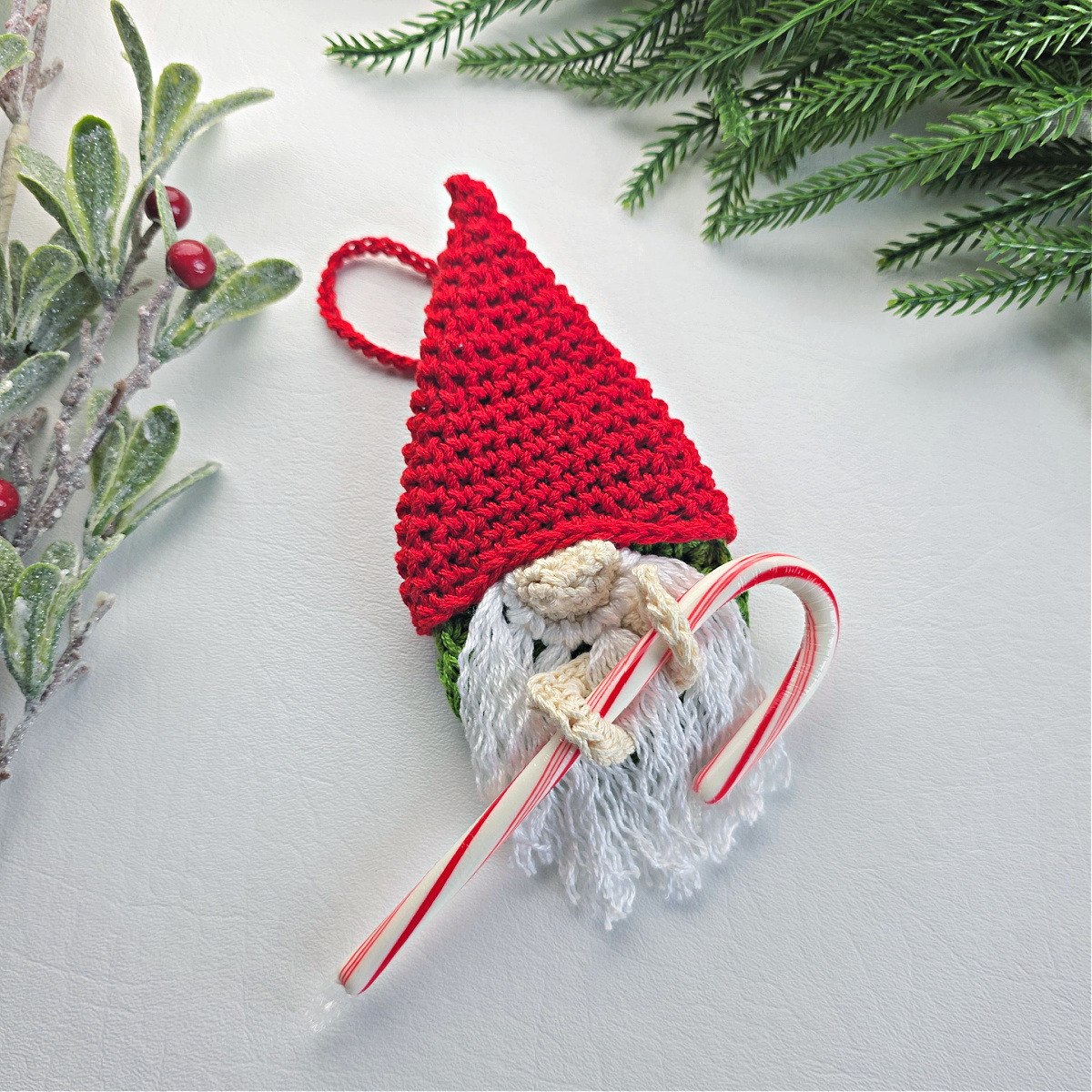 A crochet crochet gnome candy cane holder with a red santa hat next to green tinsel.