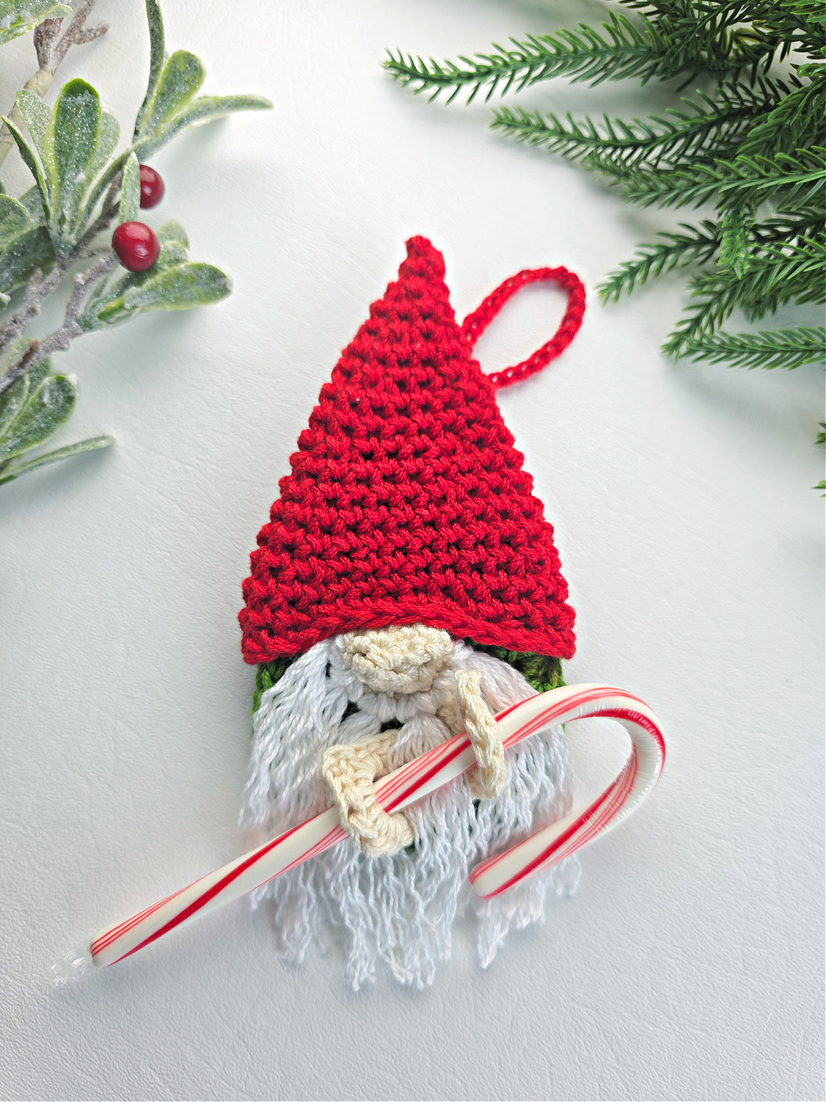 A crochet crochet gnome candy cane holder with a red santa hat next to green tinsel.