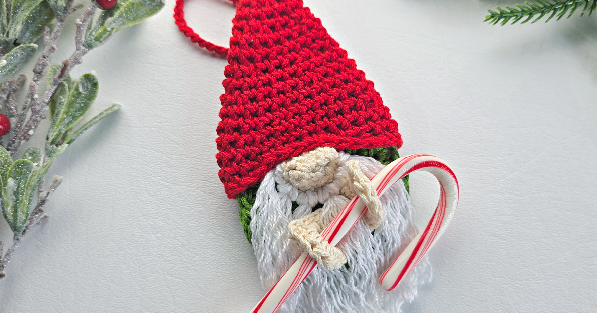 A crochet crochet gnome candy cane holder with a red santa hat next to green tinsel.