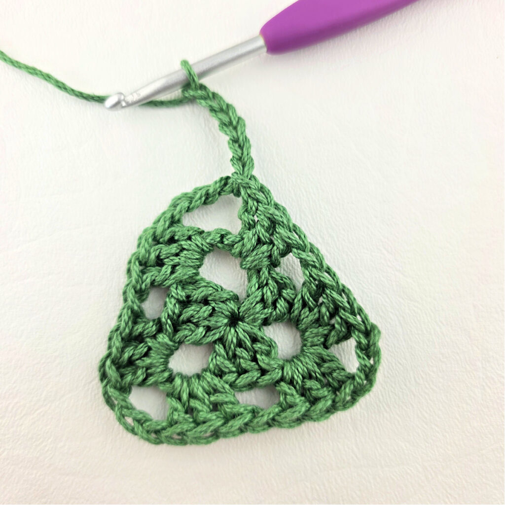 Round 3 of crochet granny stitch triangle with a purple crochet hook.
