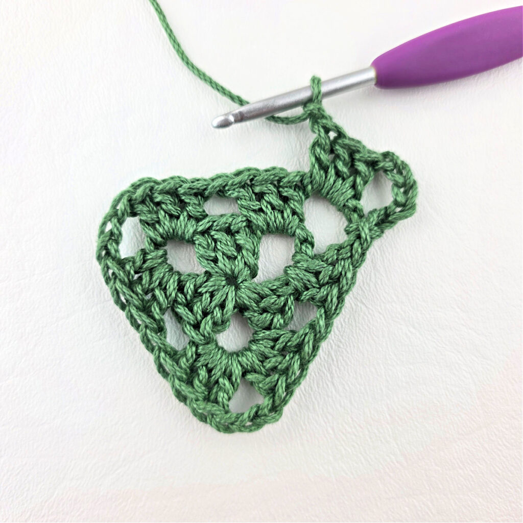 Round 3 of crochet granny stitch triangle with a purple crochet hook.