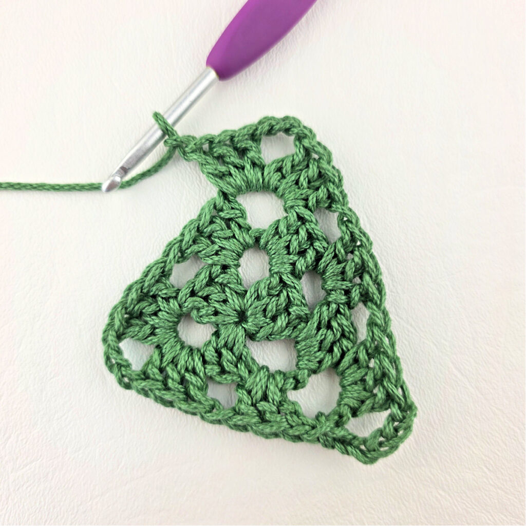 Round 3 of crochet granny stitch triangle with a purple crochet hook.