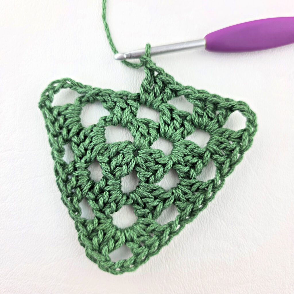 Round 3 of crochet granny stitch triangle with a purple crochet hook.