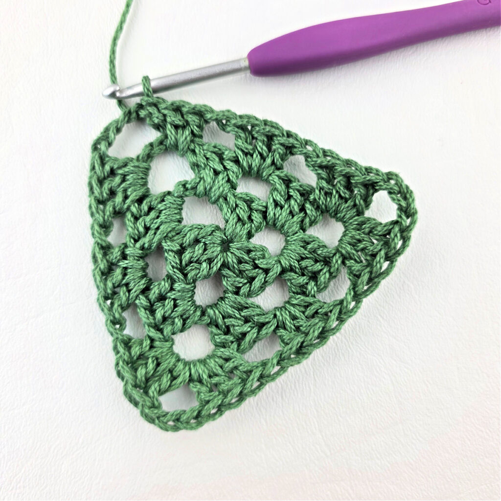 Round 3 of crochet granny stitch triangle with a purple crochet hook.