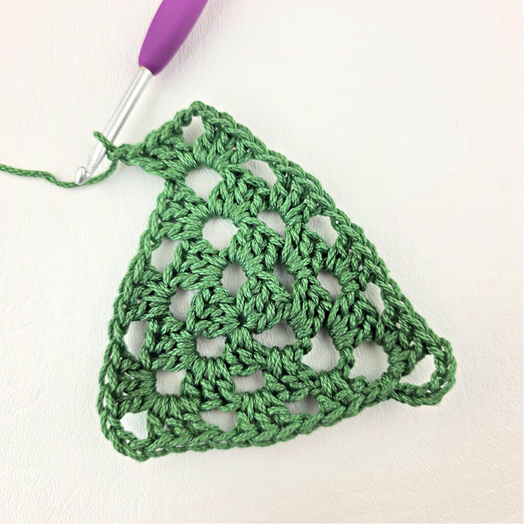 Round 4 of crochet granny stitch triangle with a purple crochet hook.
