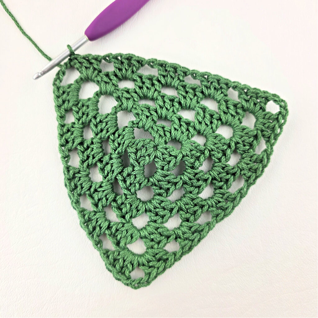 Round 5 of crochet granny stitch triangle with a purple crochet hook.